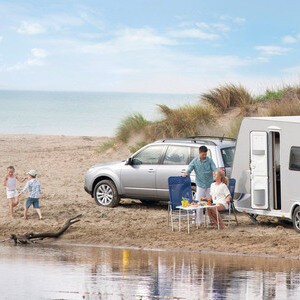 Familie mit Wohnwagen und Auto am Strand, zwei Kinder spielen am Wasser.