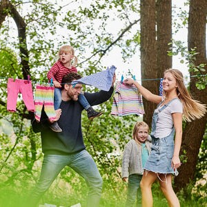 Eine lachende junge Familie hängt Wäsche auf eine Leine zwischen zwei Bäumen.