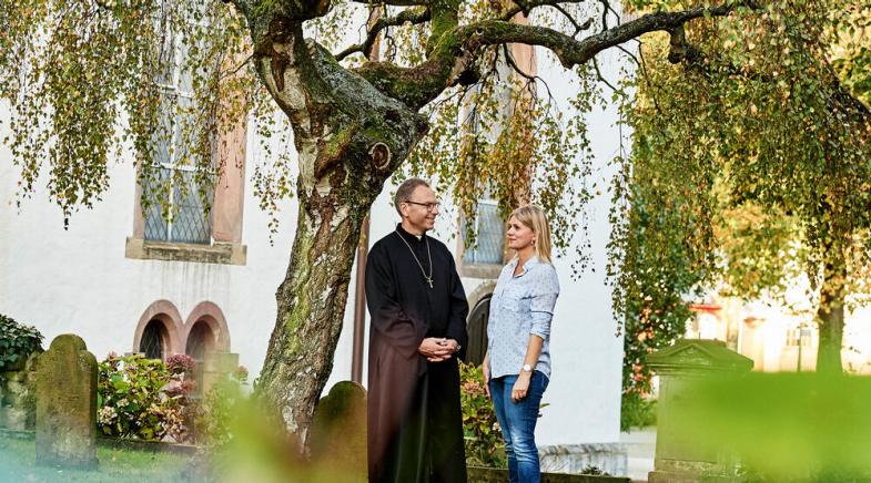 Ein Pfarrer steht mit einer Frau im Kirchengarten.