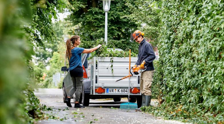 Eine Frau und ein Mann beladen einen Anhänger mit Grünschnitt.