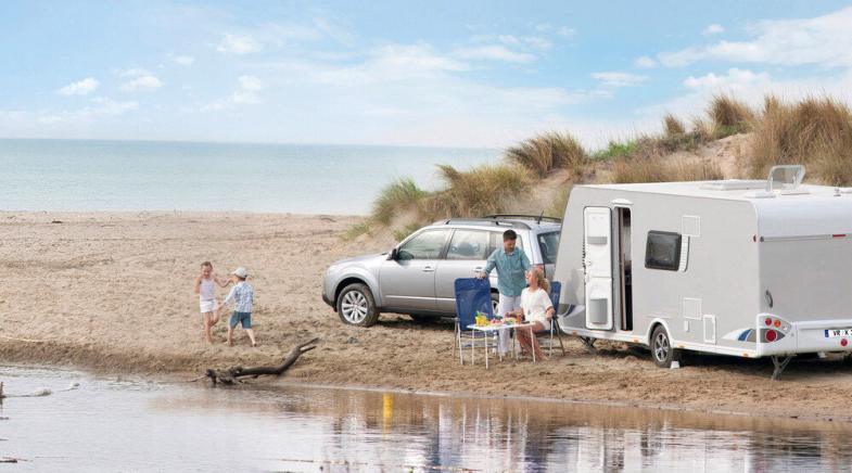 Familie mit Wohnwagen und Auto am Strand, zwei Kinder spielen am Wasser.