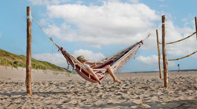 Eine Person mit Hut liegt in einer Hängematte am Strand.