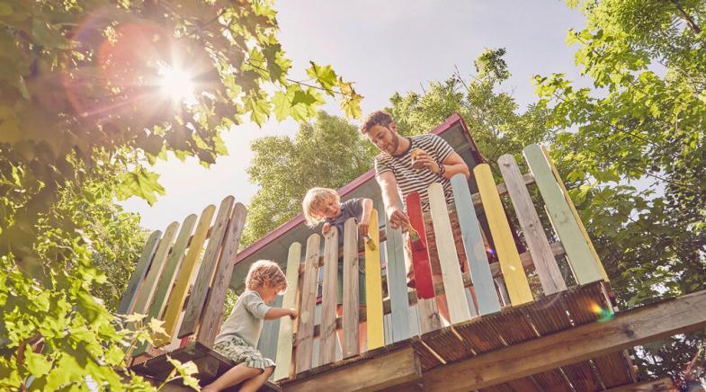 Zwei Kinder und ihr Vater streichen ein Baumhaus in bunten Farben.