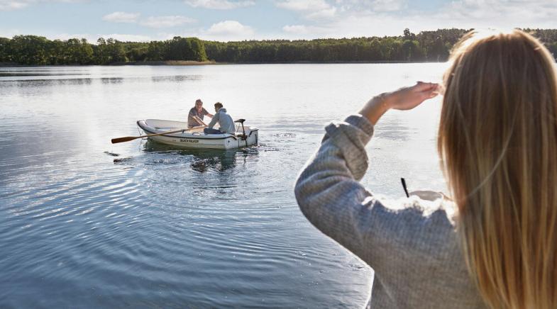 Frau schaut auf einen See. Auf dem See sitzen zwei Personen in einem Ruderboot.