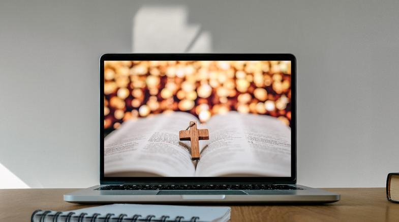 Ein Laptop auf dessen Monitor eine geöffnete Bibel mit einem darauf liegendem kleinen Kreuz aus Holz zu sehen ist.