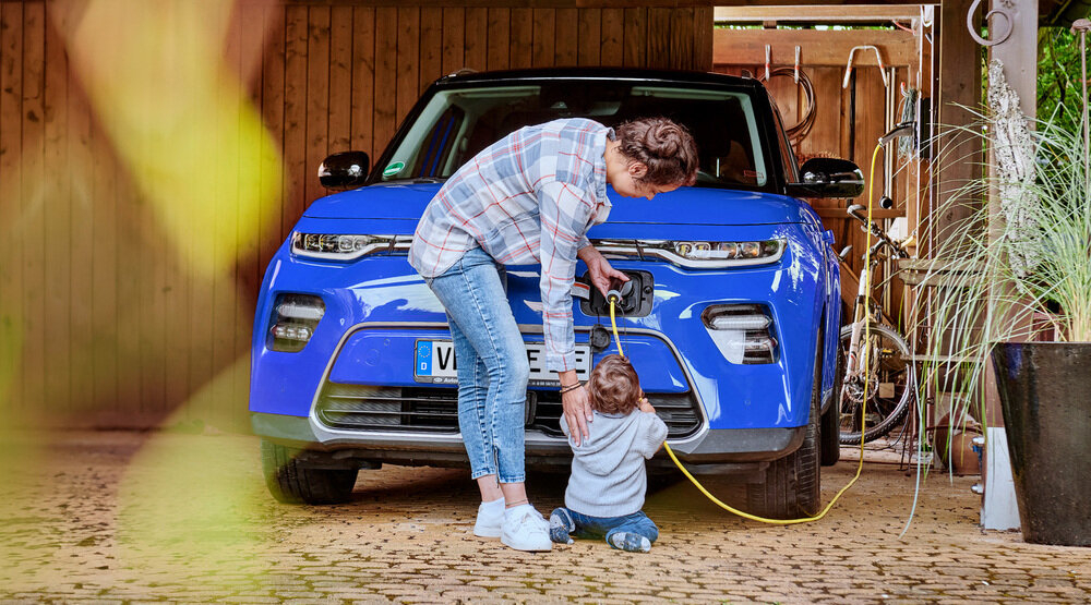 Eine Frau lädt ihr Elektroauto auf. Ihr kleines Kind hilft und hält das Kabel.