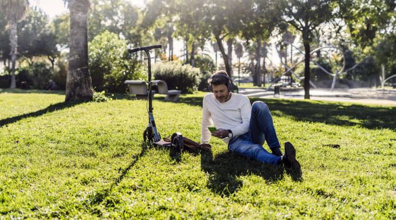 Ein Mann liegt mit Kopfhörern auf einer Wiese im Park neben seinem E-Roller.