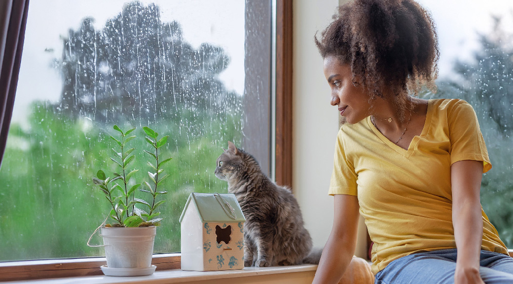 Eine junge Frau schaut mit ihrer Katze bei Regen aus dem Fenster.