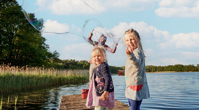 Zwei Kinder und ein Mann stehen auf einem Steg am See und machen Seifenblasen.