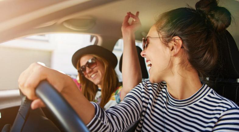 Zwei junge Frauen tragen Sonnenbrillen und singen gemeinsam im Auto.