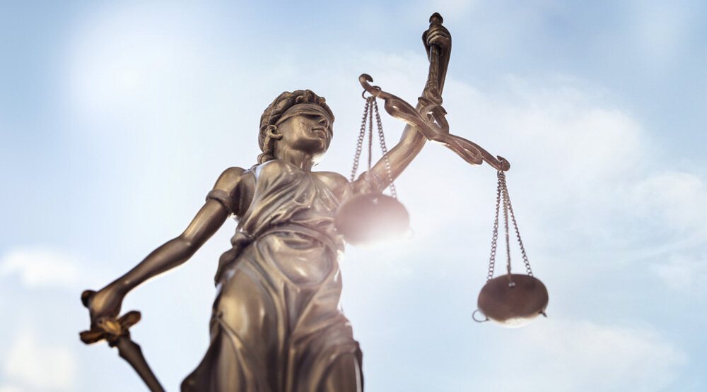 Justitia Bronzefigur vor blauem Himmel mit Wolken.