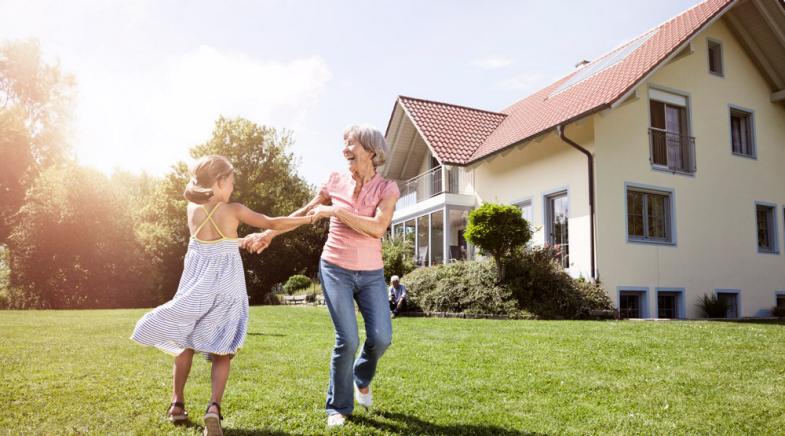 Enkelin spielt mit ihrer Oma auf einer Wiese vor einem großen Wohnhaus