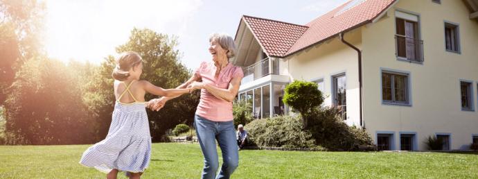 Enkelin spielt mit ihrer Oma auf einer Wiese vor einem großen Wohnhaus