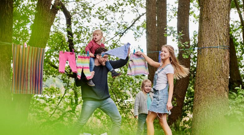 Eine lachende junge Familie hängt Wäsche auf eine Leine zwischen zwei Bäumen.