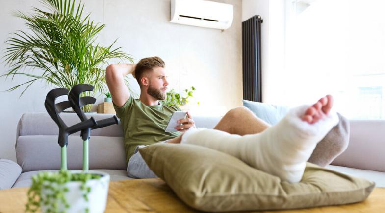 Ein junger Mann mit eingegipsten Fuß sitzt mit seinem Handy auf einer Couch.