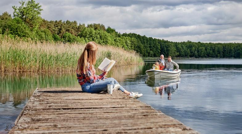 Eine Frau sitzt auf einem Steg und liest während weiterer Personen mit einem Boot auf dem See fahren.