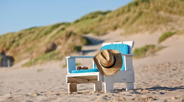 Ein Holzstuhl mit Sitzkissen, Hut und Sonnenbrille steht am Strand.