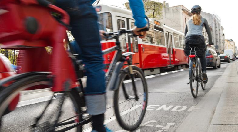 Zwei Fahrradfahrer auf dem Radweg. Es fahren eine S-Bahn und Autos neben ihnen.