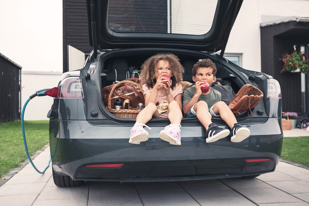 Zwei Kinder sitzen im geöffneten Kofferraum eines Autos und essen einen Apfel.