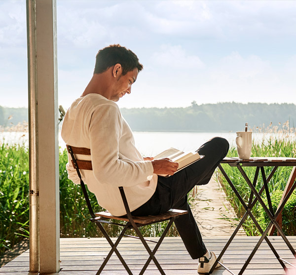 Ein Mann sitzt auf der Terrasse und liest ein Buch. Im Hintergrund liegt ein See.