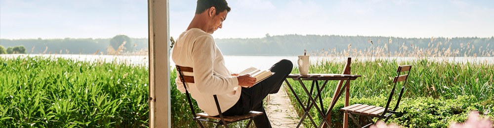 Ein Mann sitzt auf der Terrasse und liest ein Buch. Im Hintergrund liegt ein See.